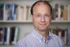 The photo shown is a portrait. Ulman Lindenberger, Director of the Center for Lifespan Psychology and Vice President of the Human Sciences Section of the Max Planck Society, is in the foreground and is pictured up to chest level. He is smiling at the camera. An out-of-focus bookshelf can be seen in the background.